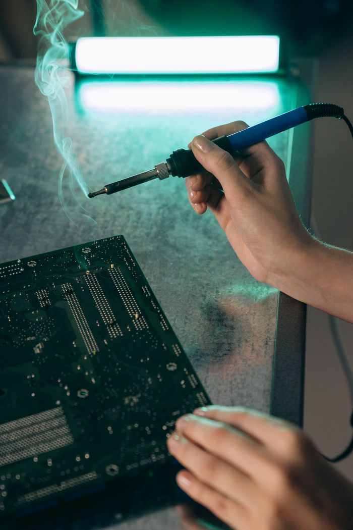 contact-img Close-up of hands working on a motherboard with a soldering iron, producing smoke.